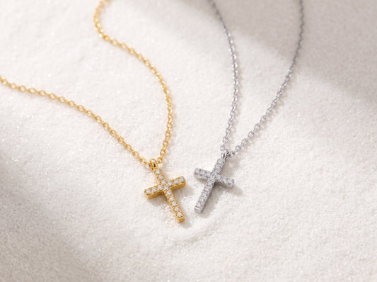 A close-up view of a dainty 14k gold-plated tiny cross necklace, featuring a delicate yellow gold chain and a petite cross pendant closely set with sparkling cubic zirconia (CZ) diamonds, shown as a fine jewelry product display and worn on a model.