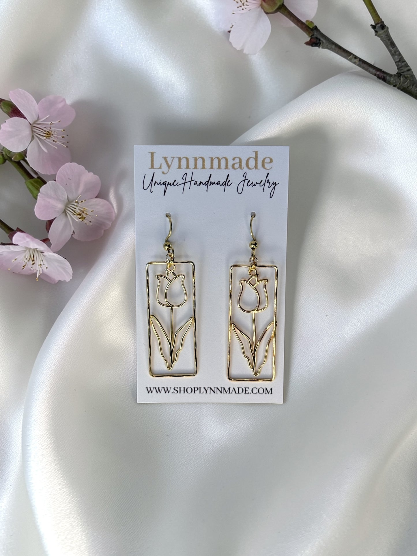 A pair of gold-toned handmade earrings featuring a minimalist wire tulip design inside a vertical rectangular frame, displayed on a white branded Lynnmade card