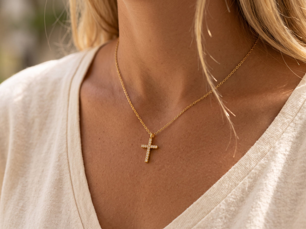 A close-up view of a dainty 14k gold-plated tiny cross necklace, featuring a delicate yellow gold chain and a petite cross pendant closely set with sparkling cubic zirconia (CZ) diamonds, shown as a fine jewelry product display and worn on a model.