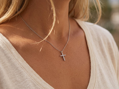A close-up view of a dainty 14k gold-plated tiny cross necklace, featuring a delicate yellow gold chain and a petite cross pendant closely set with sparkling cubic zirconia (CZ) diamonds, shown as a fine jewelry product display and worn on a model.
